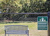 Thumbnail 9 of 46 - A sign welcomes visitors to a dog park with a bench in front of a fence.