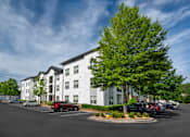 Thumbnail 2 of 57 - A parking lot in front of a white apartment building with a tree in the foreground.