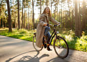 Thumbnail 8 of 11 - A woman is riding a bicycle on a paved path in a wooded area.