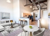 Thumbnail 1 of 31 - A modern living room with a white couch, a round table with a bowl of fruit on it, and a kitchen area in the background.