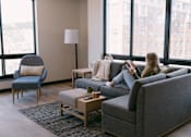 Thumbnail 58 of 73 - A woman is sitting on a grey couch in a living room with a large window behind her.