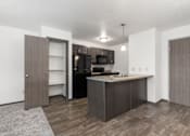 Thumbnail 12 of 31 - A kitchen with a black fridge and microwave, brown cabinets, and a grey rug.
