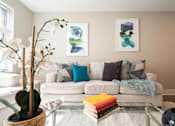 Thumbnail 28 of 50 - A living room with a white couch, a glass coffee table, and two framed artworks on the wall.