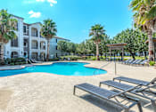 Thumbnail 25 of 36 - A swimming pool surrounded by sun loungers and palm trees in front of a multi-story apartment building at Rosemary Apartments, Conroe