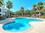 Thumbnail 31 of 36 - A large swimming pool surrounded by palm trees and a white building at Rosemary Apartments, Conroe, Texas
