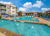 Thumbnail 5 of 27 - a swimming pool with blue lounge chairs and umbrellas in front of an apartment building at The Landings at Brooks City-Base, Texas