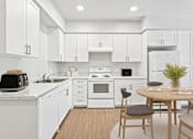 Thumbnail 6 of 29 - A modern kitchen with white cabinets and appliances, a wooden table with chairs, and a plant on the table.