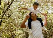 Thumbnail 1 of 9 - A man lifts a child into the air in a forest.
