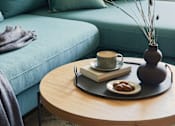 Thumbnail 2 of 9 - A living room with a blue couch, a wooden table with a vase, a book, a cup, and some fruit.