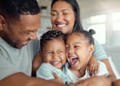 Thumbnail 3 of 9 - A family of four is hugging and smiling together in a bright room.