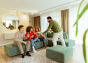 Thumbnail 4 of 18 - A man is serving food to two people sitting on a green couch in a bright room.