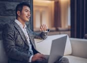 Thumbnail 7 of 9 - A man in a grey suit is sitting on a white couch using a laptop.
