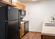 Thumbnail 11 of 18 - A black refrigerator stands in a kitchen with wooden cabinets and a stainless steel sink. Fargo, ND Cedars 4 Apartments