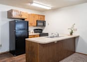 Thumbnail 10 of 18 - A kitchen with a black fridge, microwave, and wooden cabinets. Fargo, ND Cedars 4 Apartments