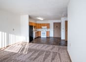 Thumbnail 16 of 21 - A sunlit living room with a carpeted floor and a kitchen area in the background. Dilworth, MN Fieldstone Apartments