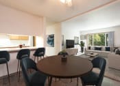 Thumbnail 2 of 7 - Bismarck, ND Bradbury Apartments. The room features a four-chair dining table with bar stools and a glimpse of the living room in the background. Soft lighting creates a warm and inviting atmosphere, while a window brings in natural light. It's a comfortable space for dining and relaxing with a seamless connection to the living room area