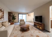 Thumbnail 1 of 10 - Bismarck, ND Newgate Apartments. A living room with a large rug, couch, and tv. A glass sliding door brightens the room with natural light