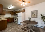 Thumbnail 2 of 17 - Bismarck, ND Rosser Apartments. A dining room with a table and two chairs. The overhead fan and light brighten the room. A kitchen with a white refrigerator freezer next to a white stove top oven are in the background