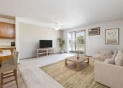 Thumbnail 1 of 16 - Sierra Ridge Apartments in Bismarck, ND | a living room with white walls and a sliding glass door. Theres a white couch, coffee table, tv and bar stools at the counter in the dining area.