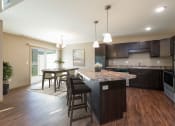 Thumbnail 4 of 11 - Bismarck, ND Stonefield Townhomes. A kitchen with dark wood cabinets and a large island with a granite countertop. The space features a well-appointed dining table, modern kitchen appliances, and tasteful decor. Bright natural light fills the room from the glass sliding door.