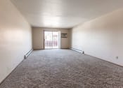 Thumbnail 5 of 19 - Grand Forks, ND Cherry Creek Apartments. A vacant living room in the unit 223 with a glass sliding door and white walls with a carpeted floor.