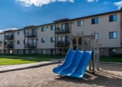 Thumbnail 30 of 32 - Fargo, ND Prairie Park Apartments. A playground with two blue slides in front of an apartment building