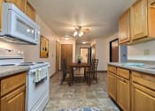 Thumbnail 5 of 21 - Fargo, ND Prairiewood Courts Apartments. A kitchen with white appliances and wooden cabinets. The dining room and table are in the background with four chairs and a light/ fan above it.