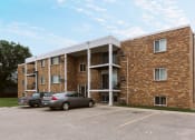 Thumbnail 2 of 2 - Grand Forks, ND Hoven Lane Apartments. A brick apartment building with two cars parked in a parking lot