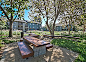 Thumbnail 14 of 39 - A wooden bench sits in the middle of a concrete area with trees and apartment buildings in the background.