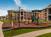 Thumbnail 10 of 16 - A playground area with a green slide and a brown sandbox in front of apartment buildings.