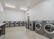 Thumbnail 13 of 16 - A row of front-loading washing machines are lined up in a laundromat.