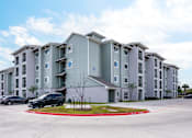 Thumbnail 7 of 9 - A large apartment complex with a parking lot in front.