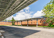 Thumbnail 23 of 39 - A long building with a metal roof and a parking lot in front.