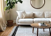Thumbnail 13 of 16 - A living room with a white couch, a round mirror, a potted plant, and a coffee table.