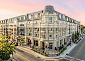 Thumbnail 1 of 35 - A large, multi-story building with a curved roof and balconies is situated on a street corner with a sidewalk and street lamps.
