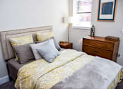 Thumbnail 5 of 7 - A bed with a yellow comforter and grey pillows sits in a room with a wooden nightstand and a small table beside it.