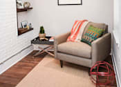 Thumbnail 6 of 7 - A living room with a grey couch, a small table, and a framed picture on the wall.