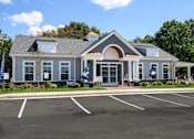 Thumbnail 2 of 35 - A building with a grey roof and white walls with a parking lot in front at The Promenade at Easton Apartments, Easton, 21601