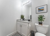 Thumbnail 20 of 28 - A bathroom with a white toilet, a white sink, a white cabinet, a white wall, a black trash can, a potted plant, and a framed picture.