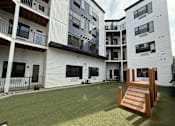 Thumbnail 23 of 26 - A wooden chair is in the foreground of a grassy area in front of apartment buildings at The Hive Apartments, Allentown, PA