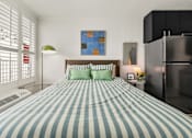 Thumbnail 3 of 20 - A bed with a blue and white striped comforter sits in a room with a painting on the wall and a refrigerator to the right.