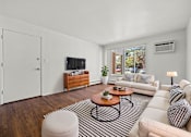 Thumbnail 2 of 26 - A living room with a white couch, a wooden coffee table, and a flat-screen TV on a wooden cabinet.
