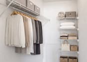 Thumbnail 10 of 24 - A white closet with clothes hanging and shelves with baskets and towels.