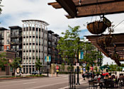 Thumbnail 22 of 22 - A street view of a city with a building in the background and people sitting at tables under a canopy.