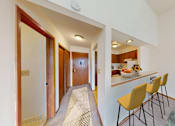 Thumbnail 3 of 10 - A hallway with a striped carpet leads to a kitchen area with a bar and chairs.