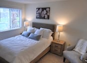 Thumbnail 42 of 43 - A bedroom with a large bed, a nightstand with a lamp, and a chair with a blanket on it.