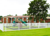 Thumbnail 30 of 61 - a white fence in front of a brick building with green grass and trees