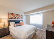 Thumbnail 7 of 18 - A bedroom with a bed, nightstand, lamp, and a painting on the wall.