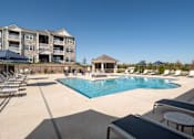 Thumbnail 7 of 36 - A large swimming pool surrounded by lounge chairs and umbrellas in front of a multi-story apartment building.
