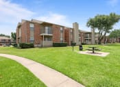 Thumbnail 30 of 47 - a picnic table sits in the middle of a grassy area in front of an apartment building at The Hudson, North Richland Hills, 76180